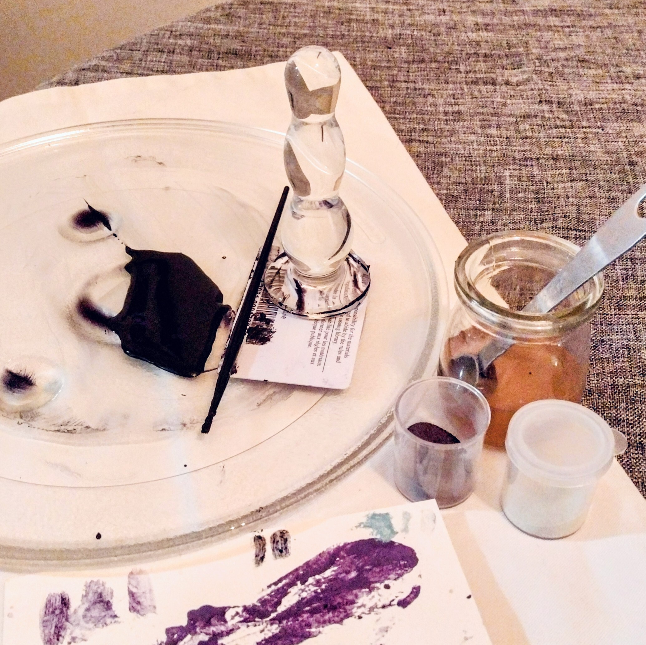 A glass microwave tray on a table with a small, dark pool of freshly mixed paint next to a brush, a library card and a glass buttplug. Next to the microwave tray are two small plastic containers, one with purple powder, and a small jar with a metal spoon in resin-coloured liquid. A card at the bottom is covered in test-streaks of different coloured paints.